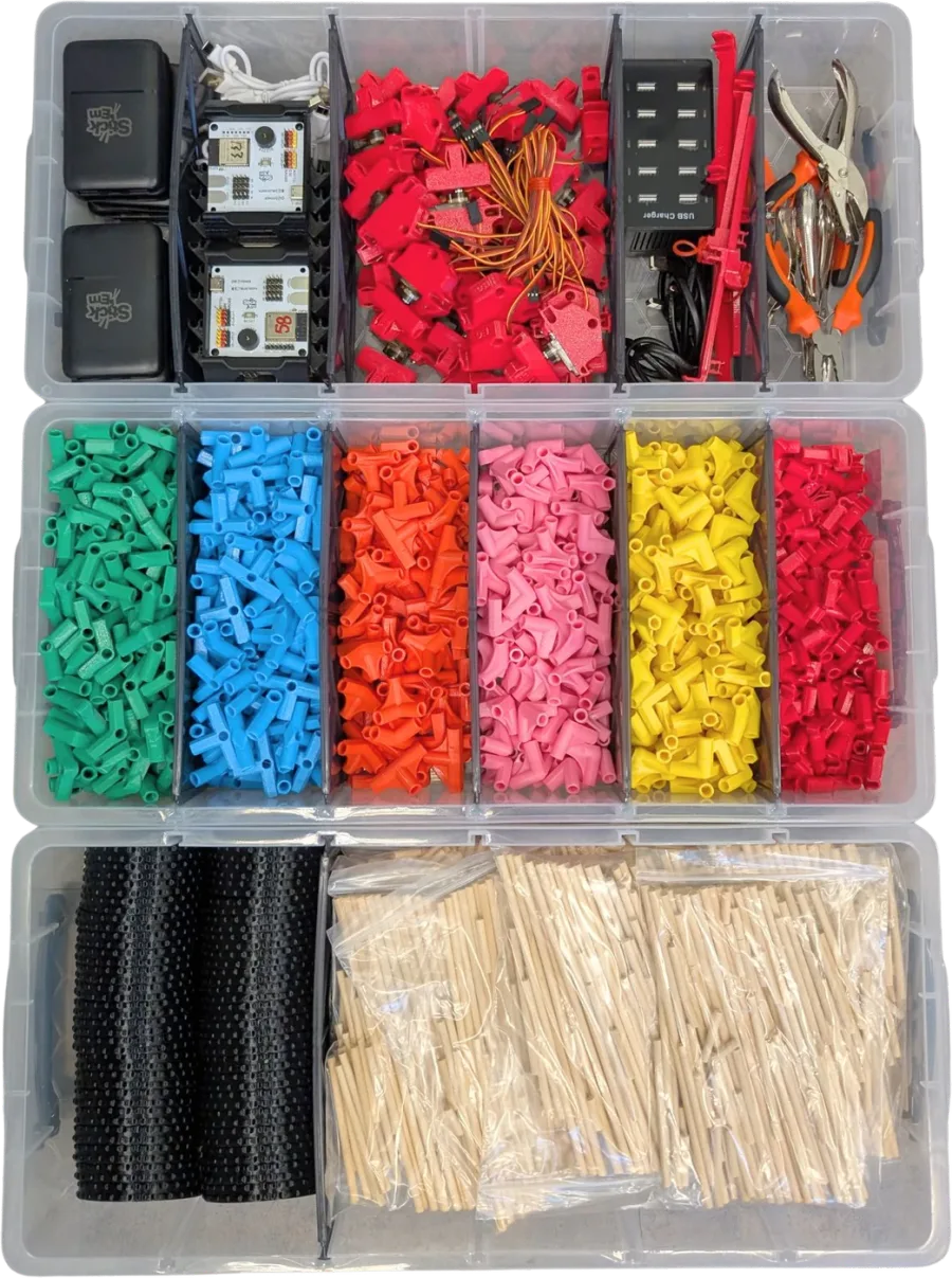 An open Stick ’Em Classroom Set in three clear trays: power banks, electronics boards, red servo motors with wiring, a USB charging hub and hand tools on top; six compartments of green, blue, orange, pink, yellow and red 3D-printed connectors in the middle; stacked black parts and bagged bamboo chopsticks at the bottom